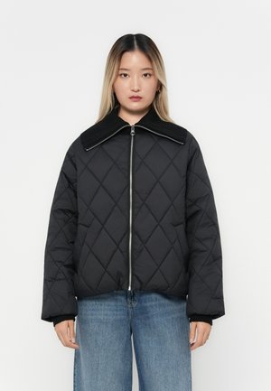 Svart quiltet jakke med bred ribbestrikket krage, glidelås foran, og lange ermer. Brukt over lyse denimjeans.