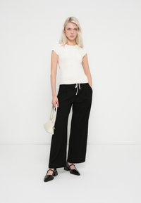 Top blanco de manga corta con textura acanalada combinado con pantalones negros de pierna ancha; la modelo sostiene un bolso blanco pequeño y lleva zapatos planos negros con punta puntiaguda.