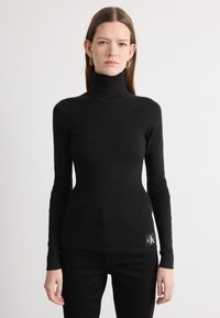 Fekete garbó pulóver bordázott textúrával, testhez álló fazonnal, hosszú ujjakkal, és egy Calvin Klein logóval a csípőn.