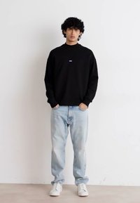 Zwarte sweatshirt met ronde hals, minimalistisch blauw logo, gecombineerd met lichtblauwe spijkerbroek en witte sneakers, staand tegen een neutrale achtergrond.