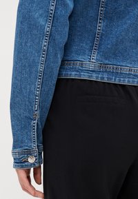 Denimjakke med blå vask, med et nært bilde av ermene som viser sømmer, en knappdetalj og en tettsittende passform. Svarte bukser synlige.