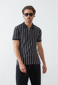 Polo noir à manches courtes avec des rayures verticales blanches, doté d'un col zippé et d'une coupe ajustée. Porté avec un pantalon sombre et des lunettes de soleil.
