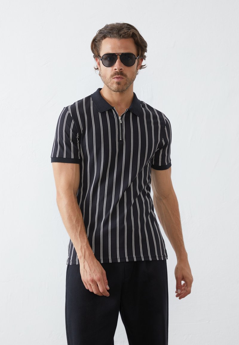 Polo noir à manches courtes avec des rayures verticales blanches, doté d'un col zippé et d'une coupe ajustée. Porté avec un pantalon sombre et des lunettes de soleil.