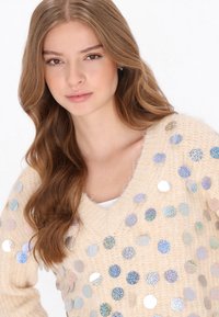 Pullover beige in maglia con scollo a V, adornato da paillettes olografiche di varie dimensioni, caratterizzato da un tessuto morbido e dalla texture lavorata.
