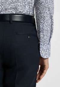 Donkerblauwe op maat gemaakte broek met een gestructureerde weving, voorzien van een achterzak met een knoop. Gekleed met een lichtbloemige overhemd.