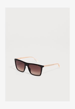 Zwarte zonnebril met vierkante montuur, bruine glazen met verloop en dunne gouden metalen pootjes, liggend op een wit oppervlak.