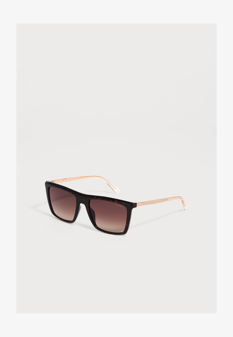 Lunettes de soleil noires à monture carrée avec verres marron dégradés et branches en métal doré fin, reposant sur une surface blanche.