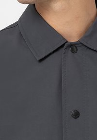 Grå, knappad skjorta med krage, som har en mjuk textur och en ensam svart knapp. Närbild som betonar sömnadsdetaljer.