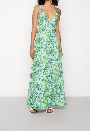 Kobieta ubrana w maxi sukienkę bez rękawów z dekoltem w serek, ozdobioną zielono-niebieskim tropikalnym liściastym wzorem, nosząca białe sandały.