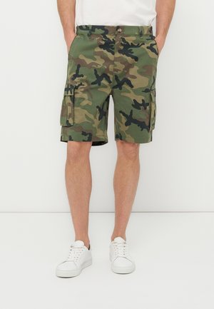 Mann trägt grüne und braune Tarn-Cargoshorts mit Seitentaschen und weiße Sneakers, steht vor einem schlichten weißen Hintergrund.