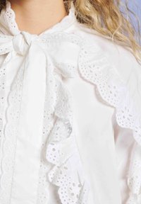 Blusa branca com rendas em ilhós e babados, com um laço atado no decote, usada com cabelo louro longo e encaracolado visível.