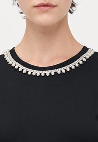 Svart topp med rund halsutskjæring prydet med en dekorativ rad av perler og rhinesteiner, som skaper en kontrast mot stoffet.