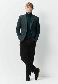 Young man standing against white wall wearing a dark green turtleneck, grey blazer, brown corduroy pants, and black shoes.