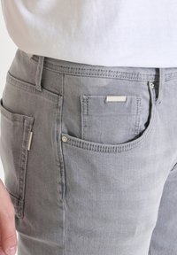 Vaqueros de mezclilla gris con un corte clásico, bolsillos delanteros y traseros, detalles metálicos plateados y una textura sutil.
