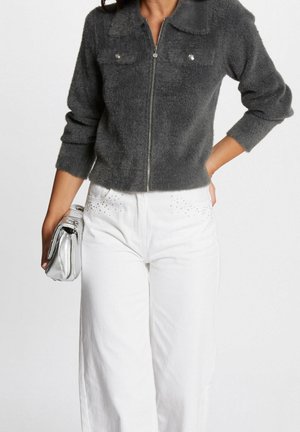 Kvinde iført en grå, blød lynlåstrøje, hvide højtaljede bukser med små nitter, holdende en sølvfarvet clutch-taske.