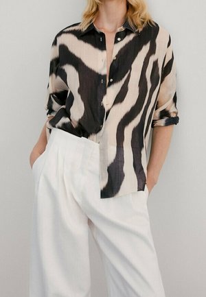 Woman wearing a black and beige abstract patterned button-up shirt with rolled sleeves and high-waisted loose white pants, hands in pockets.