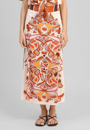 Femme portant une longue jupe avec des motifs floraux abstraits orange, rose et marron, associée à des sandales noires à brides.