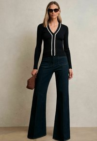 Fekete bordázott, hosszú ujjú felső, fehér díszítéssel, amely magas derekú sötét farmerral kombinálva van. Kiegészítve egy barna szőtt táskával.