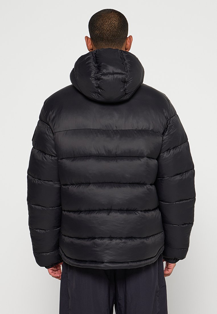 Doudoune noire avec un design texturé en matelassage. Dotée d'une capuche et d'une fermeture éclair, elle offre une coupe ample avec des accents de couture visibles.