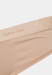 Tanga bege da Calvin Klein com uma textura suave e em relevo, e uma cintura fina com o logótipo da marca em relevo. Design monocromático e sem costuras.