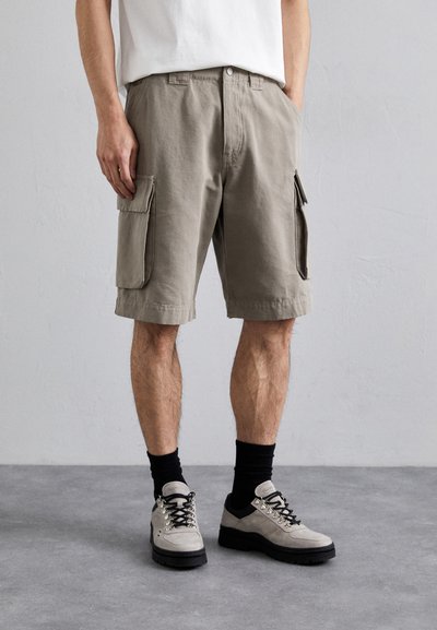 Pantaloni scurți cargo de culoare bej deschis, din bumbac, cu două buzunare mari pe lateral, purtați cu șosete negre până la gleznă și pantofi de drumeție gri.