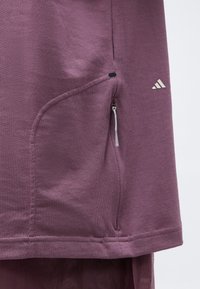 Gros plan sur un tissu violet montrant une poche latérale zippée et un petit logo Adidas blanc sur un vêtement.
