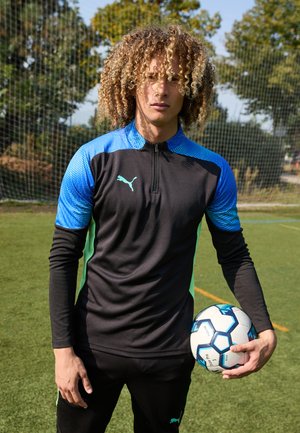 Zwarte sporttop met lange mouwen en blauw patroon op de schouders, PUMA-logo op de borst, gecombineerd met zwarte broek; houder van een witte en blauwe voetbal.