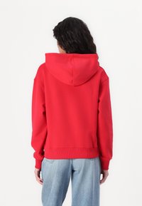 Rode katoenen capuchontrui met geribde boorden bij de mouwen en zoom, met een ontspannen pasvorm en een grote capuchon aan de achterkant. Draagt bij een blauwe spijkerbroek.