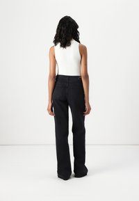 Nainen, jolla on kihara tumma tukka, pukeutuneena valkoiseen hihattomaan toppiin ja löysiin mustiin farkkuihin, seisoo yksinkertaista valkoista taustaa vasten.