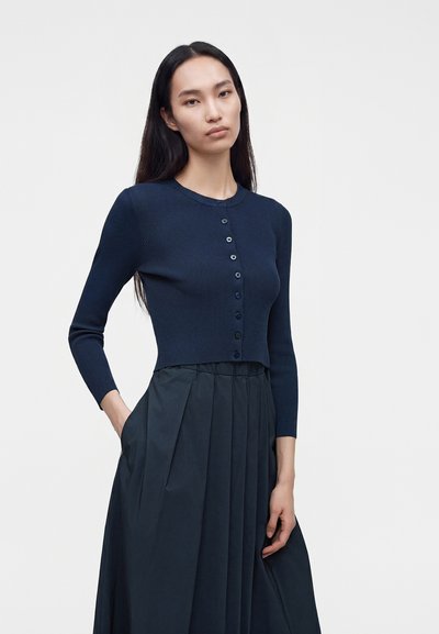 Femme aux longs cheveux noirs portant un cardigan ajusté bleu marine boutonné et une jupe plissée taille haute bleu marine, debout devant un fond uni.