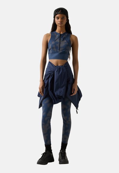 Ung kvinde iført en marineblå sports-bh, matchende camouflage-leggings, sorte sneakers, en jakke bundet om taljen og en sort kasket vendt bagud.