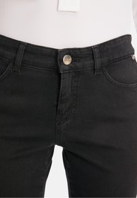 Gros plan sur une personne portant un jean en denim noir avec un bouton argenté et des passants pour ceinture, associé à une chemise blanche rentrée.