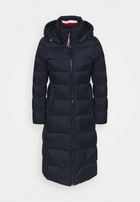 Donkerblauwe pufferjas met een capuchon, voorzien van een ritssluiting, een gestikte ontwerp en licht opgezette mouwen. De binnenkant van de kraag heeft rode en witte strepen.