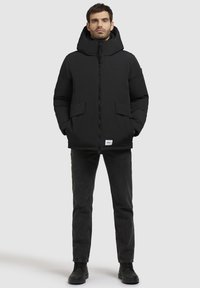 Chaqueta de plumas negra con un cuello alto y capucha, que presenta un cierre frontal y dos grandes bolsillos laterales. Llevada sobre jeans y zapatos negros.