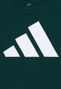 Tumši zaļš kokvilnas T-krekls ar lielu baltu trīs līniju Adidas logotipu. Apaļa kakla izgriezums ar gludu tekstūru un bez papildu rakstiem.