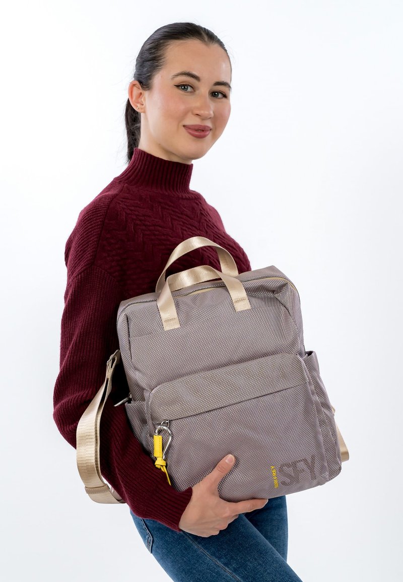 Szary plecak z materiału z elementami w złotym kolorze, wyposażony w dwie rączki i regulowany pasek. Z przodu znajduje się kieszeń oraz żółty suwak jako akcent.