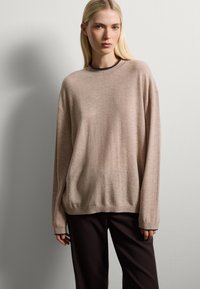 Pull oversized beige clair avec un col rond, des épaules tombantes et des poignets côtelés, associé à un pantalon marron foncé. Texture lisse.