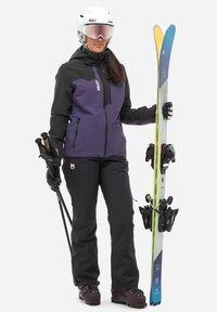 Skiausrüstung mit einer lila und schwarzen isolierten Jacke, schwarzen Hosen, Handschuhen und einem Helm. Hält farbenfrohe Skier und Skistöcke.