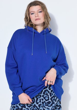 Junge Person mit welligem Haar, die einen blauen Hoodie und einen gemusterten Rock trägt, posiert mit einer Hand, die den Hoodie leicht hochzieht.
