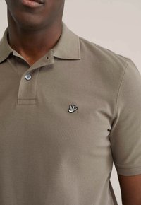 Khaki poloshirt met een gestructureerde weving, een kraag met twee knopen en een zwart geborduurd vogel-logo op de borst. Blauwe knopen zorgen voor contrast.