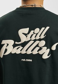 Tmavě zelené bavlněné tričko s velkým bílým kurzivním textem "Still Ballin’" a "est. 2006" v tučném, strukturovaném tisku.