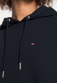 Mörkblå hoodtröja gjord av en bomullsblandning. Har metalldetaljer, dragsko och en liten logotyp i rött, vitt och blått på bröstet.