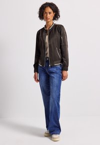 Femme debout portant une veste bomber noire sur un haut beige, un jean bleu et des baskets blanches devant un fond blanc uni.
