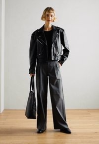 Svart skinnjakke med kort lengde og sølvfarger, kombinert med høy midje og vide grå bukser. Modellen holder en svart tote-veske.