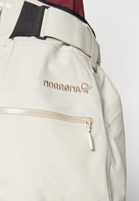 Pantalons d'extérieur beiges avec une poche zippée, un logo brodé et une ceinture robuste avec une boucle noire. Texture lisse dans l'ensemble.