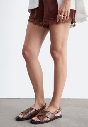 Jambes portant un short en dentelle marron et des sandales marron à motif peau de serpent avec des lanières croisées sur un sol gris avec un fond blanc.