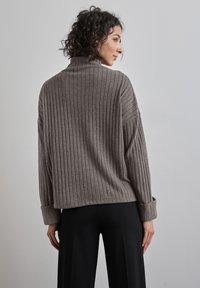 Grauer, gerippter Pullover mit hohem Kragen und weiten Ärmeln. Verfügt über umgeschlagene Bündchen und eine lockere Passform. Getragen mit schwarzen Hosen.