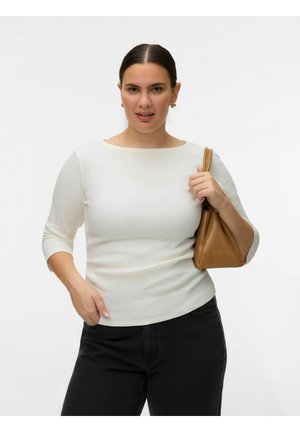 Femme aux cheveux foncés, portant un haut blanc et un pantalon noir, tenant un sac à main marron sur l'épaule, sur un fond blanc uni.