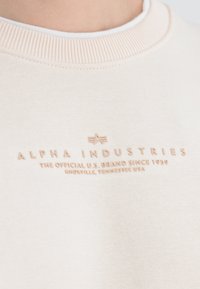 Cremfarbener Sweatshirt mit dem "Alpha Industries" Logo und dem Text "Die offizielle US-Marke seit 1959 Knoxville, Tennessee USA" auf der Brust.