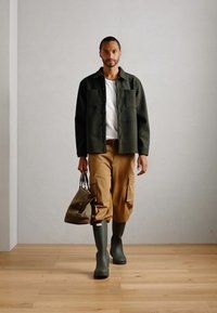 Veste chemise en mélange de laine verte sur une chemise blanche, pantalon cargo beige, bottes imperméables vert olive et un sac marron avec une poignée rayée.